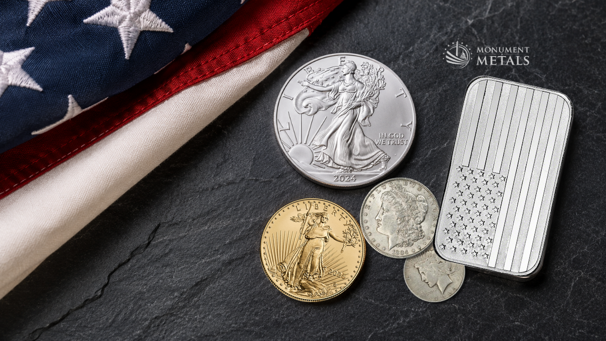 A folded American flag rests on a dark slate surface beside a curated collection of American precious metals. The display includes an American Silver Eagle, an American Gold Eagle, a Highland Mint American flag silver bar, a vintage Morgan Silver Dollar, and smaller historic U.S. silver coins arranged in a clean, premium flat lay with sharp detail and soft natural lighting.