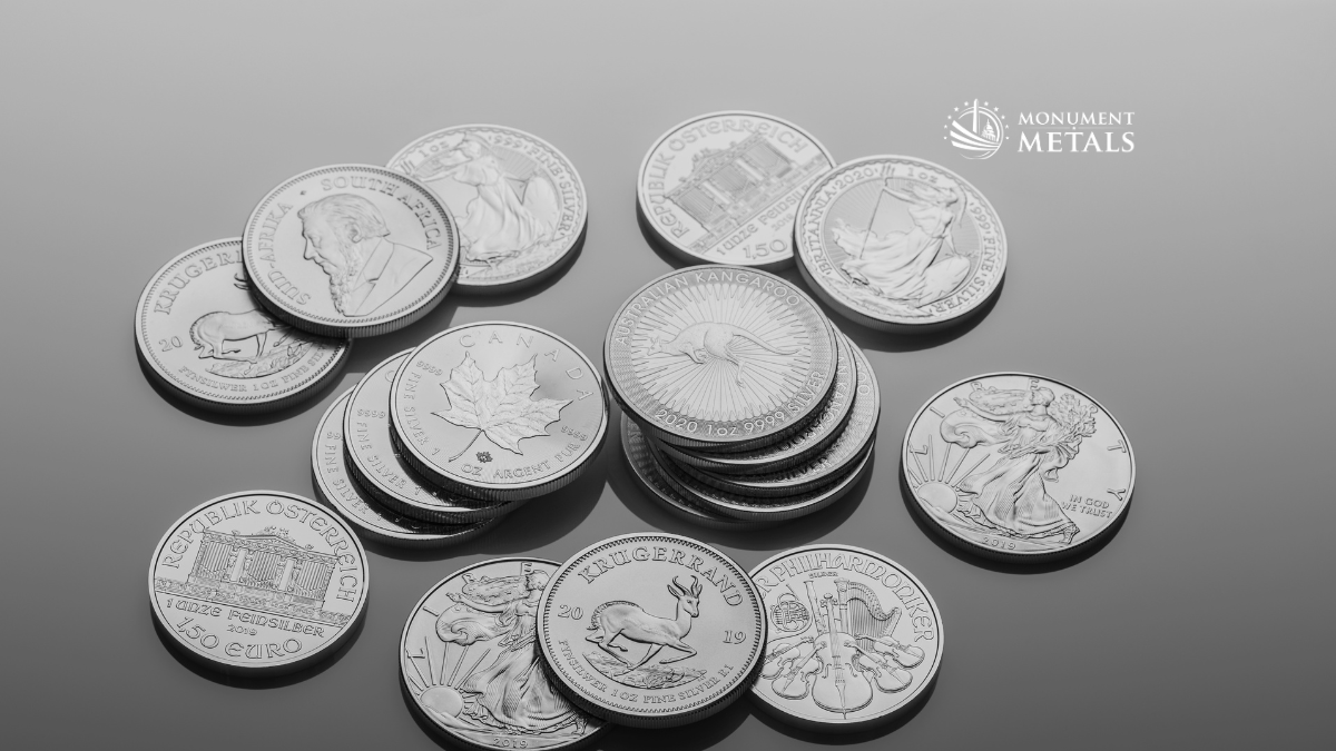 Assorted stacks and spreads of 1 oz silver bullion coins including American Silver Eagles, Canadian Maple Leafs, Austrian Philharmonics, South African Krugerrands, and Australian Kangaroos arranged on a gray studio background with Monument Metals logo.