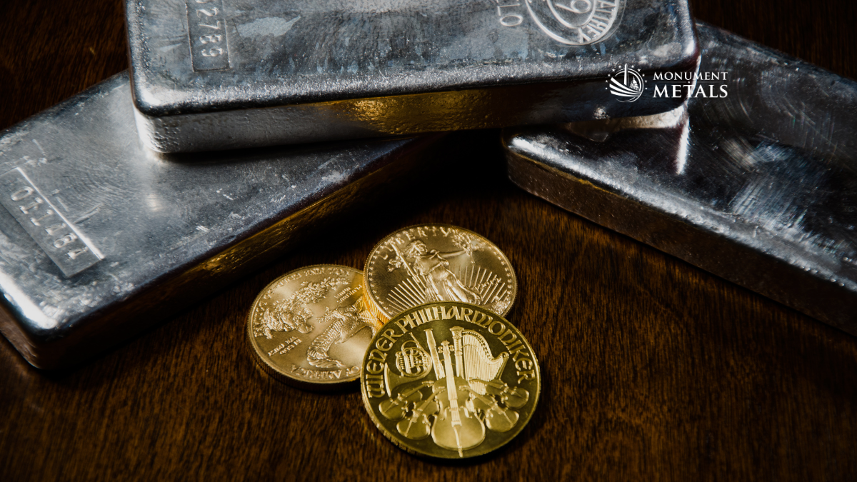 Close-up of silver bullion bars stacked on a wooden surface with gold bullion coins, including a Philharmonic and other popular designs, with the Monument Metals logo in the upper right, representing buying or selling physical precious metals.