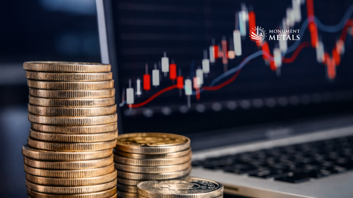 Stacks of gold coins arranged beside a laptop displaying a candlestick price chart with upward market movement, representing gold market trends and precious metals price analysis, with Monument Metals logo.
